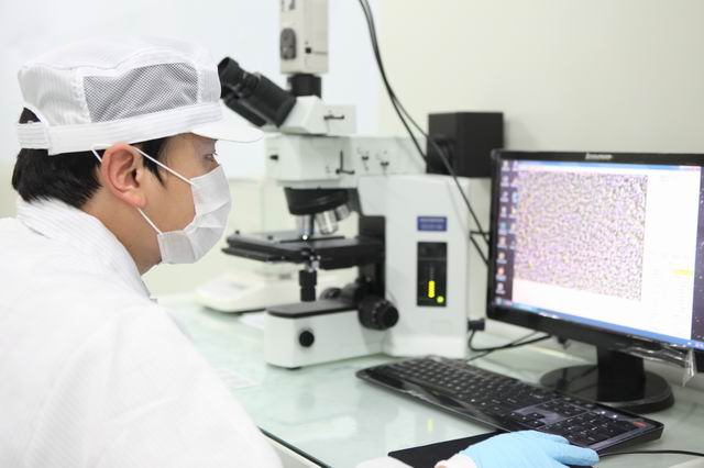 Scientist wearing protective gear and mask analyzing samples under a microscope, with results displayed on a computer screen in a laboratory setting.